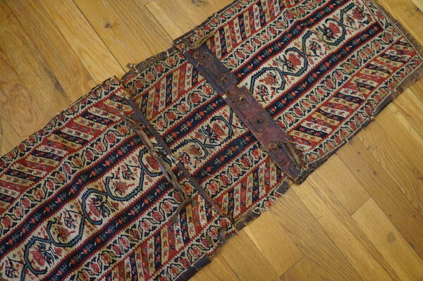 Geometric red, blue, and cream banded textile piece featuring dark leather straps on wooden floor.