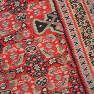 Coarse, red medium-pile rug detail featuring colorful dense patterns and layered geometric borders on wood flooring.