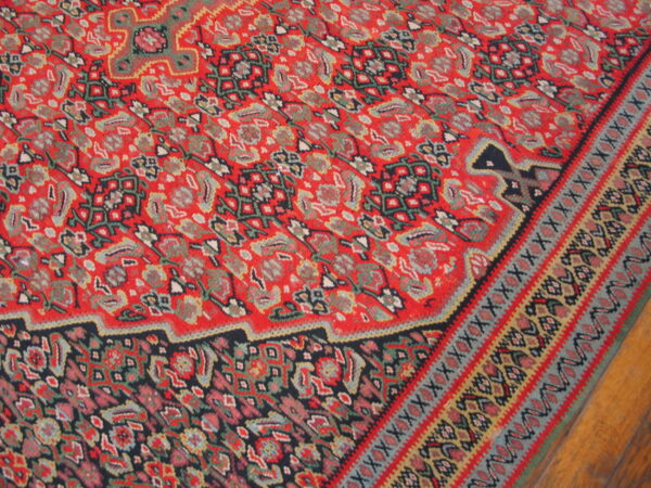 Red rug with dense, multicolored floral and geometric patterns resting on light wood flooring.
