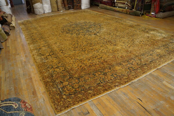 Yellow and dark blue traditional rug on worn wood floors, surrounded by stored, rolled rugs.