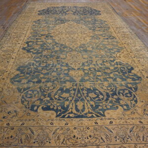 Low-pile blue and tan traditional rug with complex floral motifs rests on worn wooden floorboards.