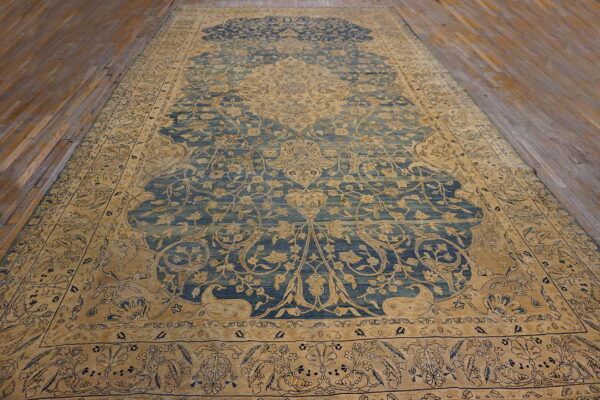Low-pile blue and tan traditional rug with complex floral motifs rests on worn wooden floorboards.