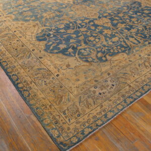 Intricate low-pile rug with a blue field and gold floral borders resting on warm wooden plank flooring.