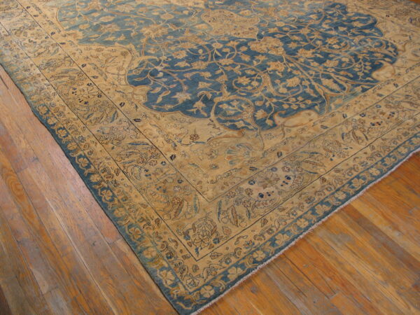 Intricate low-pile rug with a blue field and gold floral borders resting on warm wooden plank flooring.