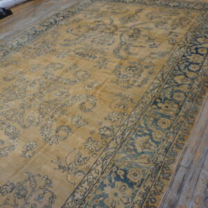 A large low-pile tan rug featuring faded blue traditional floral designs on rustic wood plank flooring.