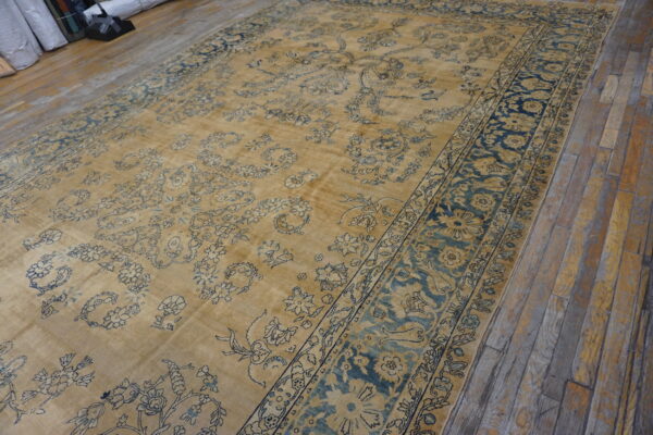 A large low-pile tan rug featuring faded blue traditional floral designs on rustic wood plank flooring.