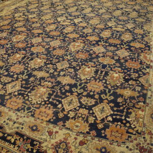 Intricate, large navy rug featuring gold and rust stylized designs and broad multi-layered beige borders.