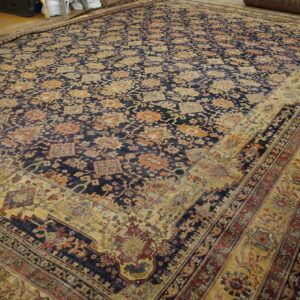 Large navy and gold floral rug with wide multiple borders laid on a wooden floor in a storage area.
