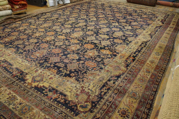 Large navy and gold floral rug with wide multiple borders laid on a wooden floor in a storage area.