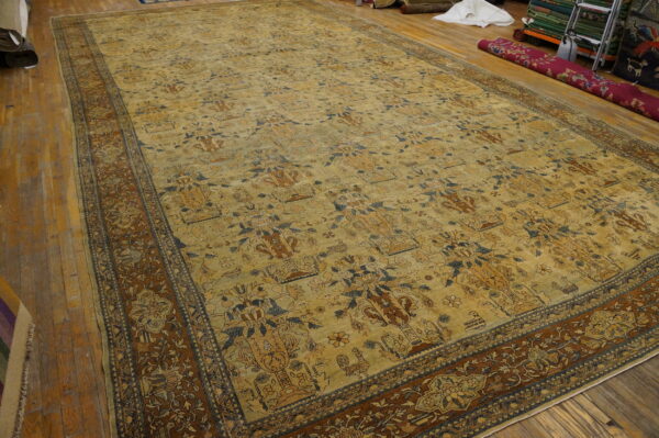 Large low-pile rug with beige field, blue and brown vase motifs, and a rust-toned floral border on wooden flooring.