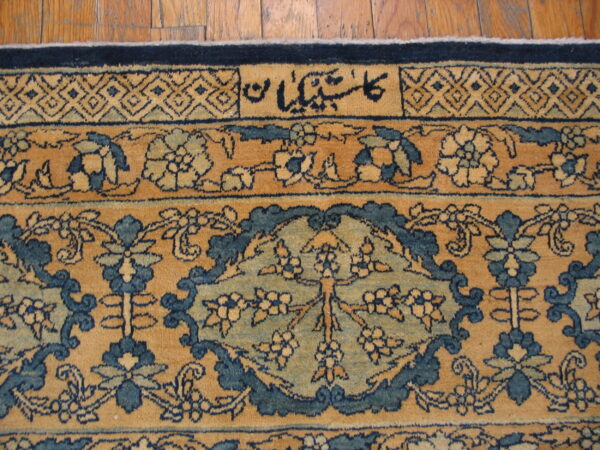 Tan and blue patterned rug detail featuring floral motifs, a geometric border, and script on wooden planks.