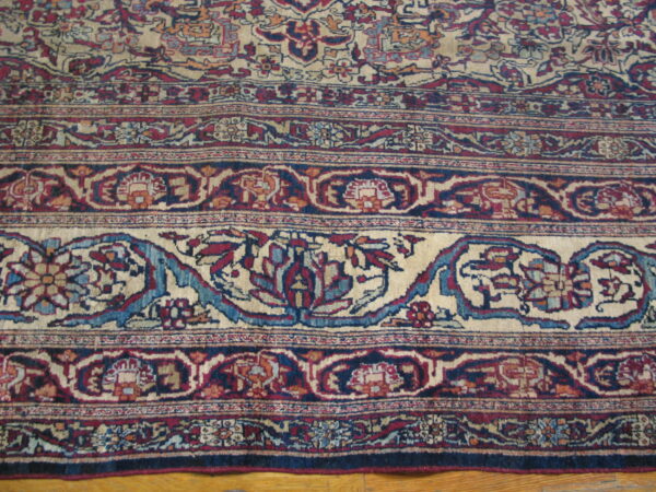 Intricate floral and vine rug border in cream, blue, and red rests on light wood planks.