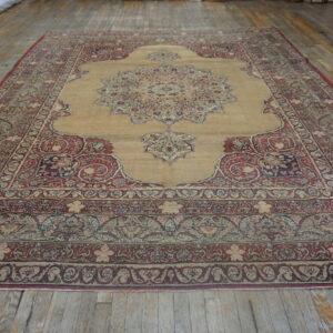 Ornate medallion rug with red borders and a tan central field displayed on aged wooden flooring.