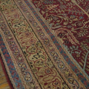 Ornate red rug with beige, pink, and blue floral borders rests on rustic light wood flooring.