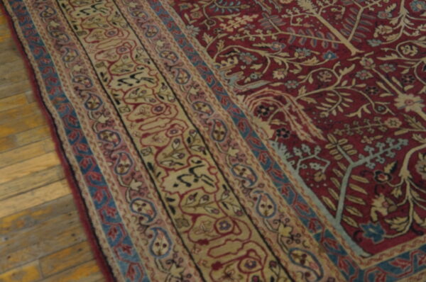 Ornate red rug with beige, pink, and blue floral borders rests on rustic light wood flooring.