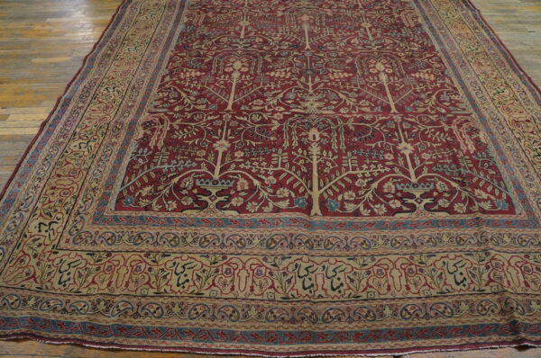 Oversized red rug with tree of life pattern and complex beige borders lying on worn wood flooring.