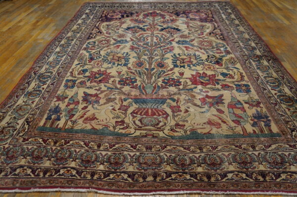 Low-pile rug with a detailed tree of life and bird pattern on a beige field, laid over wood planks.
