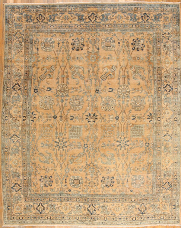 Golden tan rug with an elaborate blue and camel floral pattern and border on wood flooring.