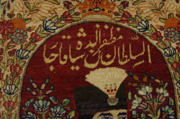 Ornate rug detail showing red calligraphy, part of a figure, and intricate cream and multicolored floral borders.