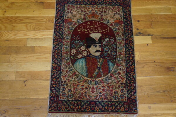 Red and blue highly detailed rug with a central portrait motif and script rests on wide plank wood flooring.
