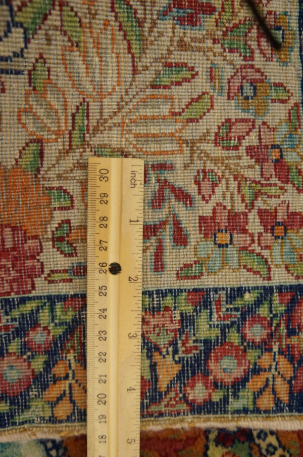 Close-up of a densely woven rug showing red and green floral motifs on a light ground, measured by a ruler.