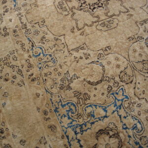Distressed tan rug with faded dark brown and bright blue traditional motifs rests on hardwood flooring.