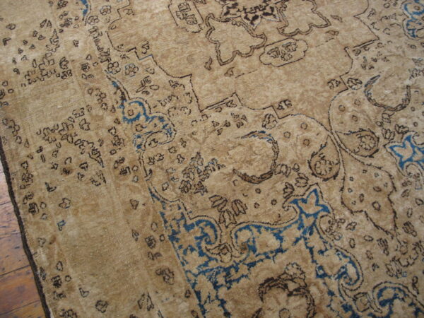 Distressed tan rug with faded dark brown and bright blue traditional motifs rests on hardwood flooring.