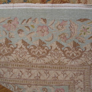 Low-pile rug detailed with muted pink flowers, tan scrollwork, and a pale blue-green field on wood flooring.