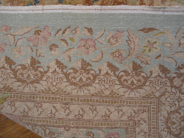 Low-pile rug detailed with muted pink flowers, tan scrollwork, and a pale blue-green field on wood flooring.