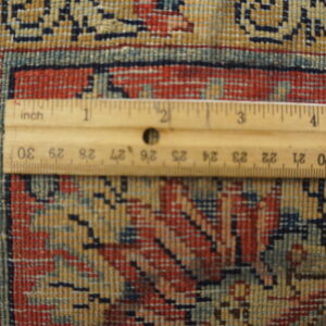 A ruler rests on the short pile rug showing the dense red, blue, and gold geometric pattern.