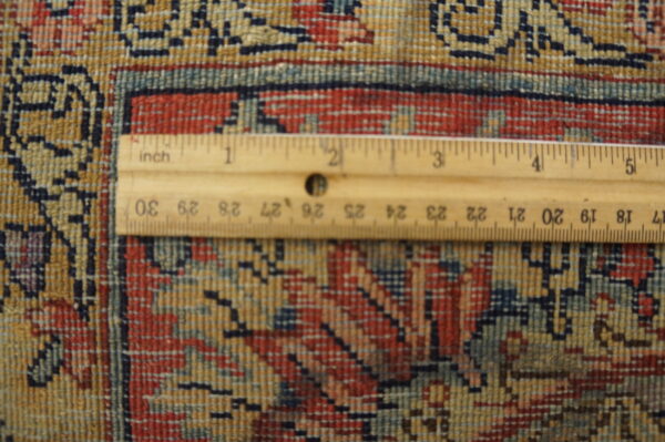A ruler rests on the short pile rug showing the dense red, blue, and gold geometric pattern.
