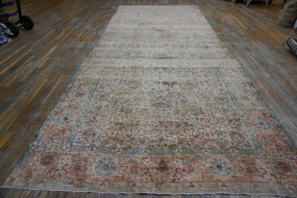 A large, distressed traditional rug in ivory, blue, and terracotta lies flat on a worn wood plank floor.