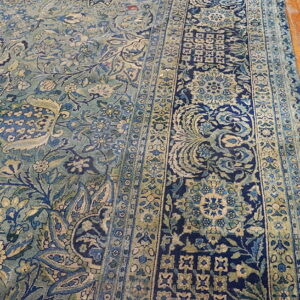 Detailed view of a blue and green low-pile rug with intricate borders on hardwood flooring.