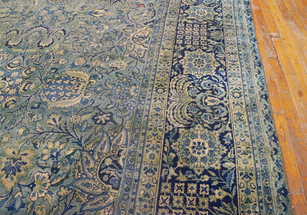 Detailed view of a blue and green low-pile rug with intricate borders on hardwood flooring.