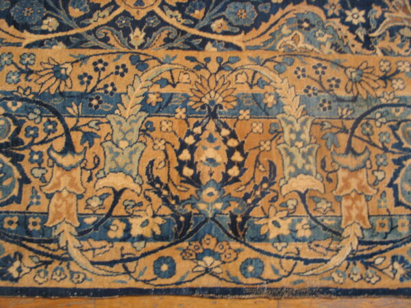 Detailed floral rug pattern in navy, teal, and golden tan rests close-up on a wooden floor.