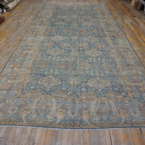 Ornate blue and gold patterned area rug displayed on worn wooden flooring with stacked rug rolls nearby.