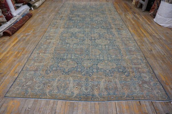 Ornate blue and gold patterned area rug displayed on worn wooden flooring with stacked rug rolls nearby.