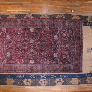 A red and blue ornate rug is displayed over a larger tan and navy patterned rug on a wood floor.