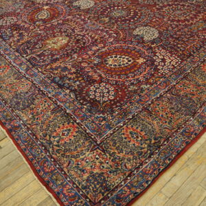 The corner of a low-pile rug featuring complex red, blue, and gold botanical patterns on distressed wooden planks.