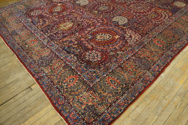The corner of a low-pile rug featuring complex red, blue, and gold botanical patterns on distressed wooden planks.