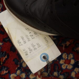 A black shoe and white inventory tag rest on a high-pile red rug with blue and tan floral patterns.