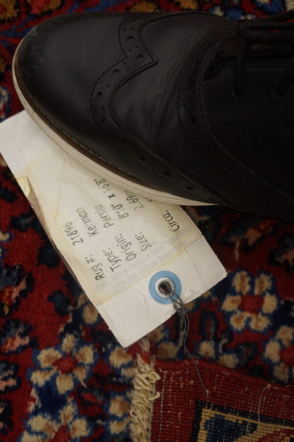 A black shoe and white inventory tag rest on a high-pile red rug with blue and tan floral patterns.