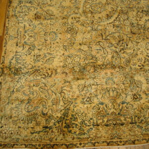 Muted golden rug with complex blue and dark brown floral motifs resting on brown hardwood planks.