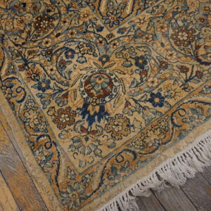 Ornate gold rug with blue and brown floral motifs, showing white fringe on rustic wooden flooring.