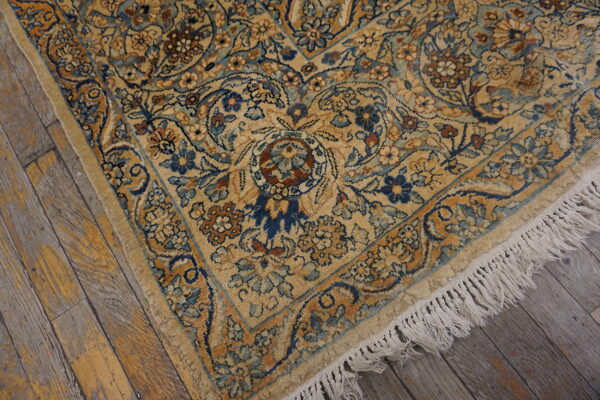 Ornate gold rug with blue and brown floral motifs, showing white fringe on rustic wooden flooring.