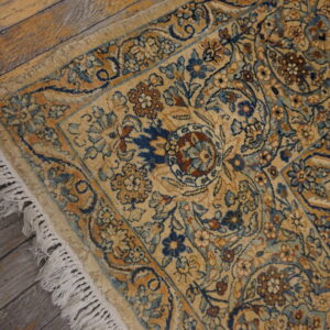 Gold rug with intricate blue and rust floral designs and fringe, set against worn wood planks.
