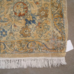 Tan floral rug detail with blue and terracotta patterns, white fringe, and a grey concrete background.