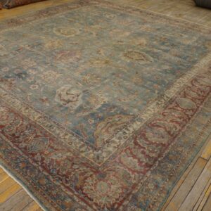 Distressed square rug with a faded teal field and wide, ornate brick red border on wood flooring.
