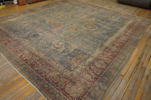 Distressed square rug with a faded teal field and wide, ornate brick red border on wood flooring.