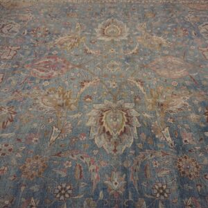 A distressed blue rug displays an intricate traditional floral and vine pattern in muted beige and rust tones.